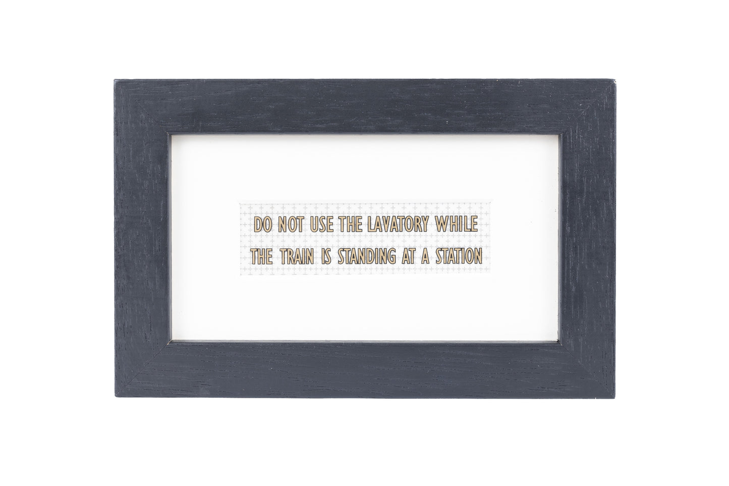 A black frame with a white background and a Do not use the lavatory while the train is standing at a station gold text on a small Formica print background