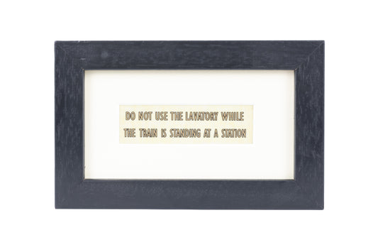 A black frame with a white background and a Do not use the lavatory while the train is standing at a station gold text on a small Formica print background