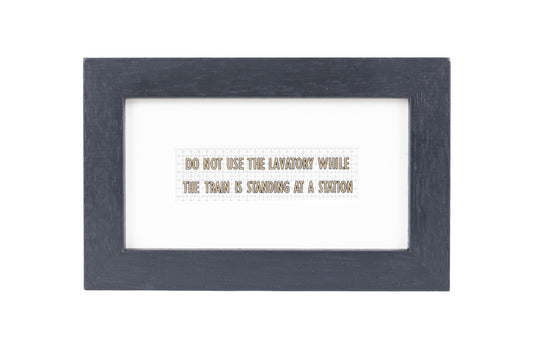 A black frame with a white background and a Do not use the lavatory while the train is standing at a station gold text on a small Formica print background
