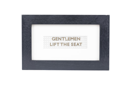 A black frame with a white background and a Gentlemen lift the seat gold text on a small Formica print background